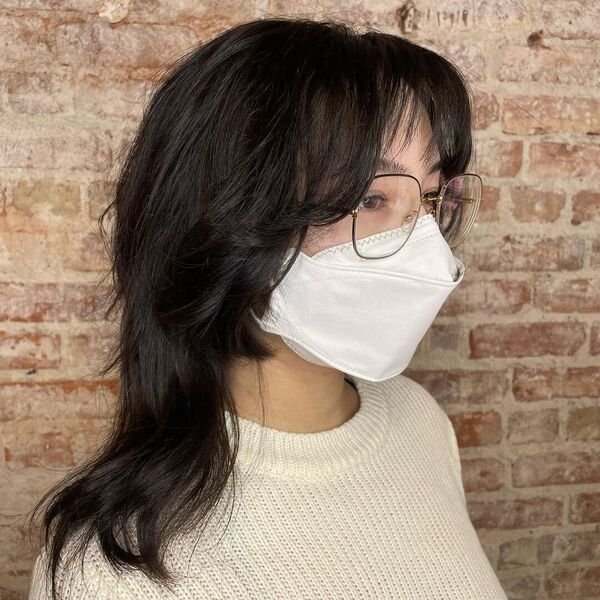 A woman wearing a knitted cream top, white mask, eyeglasses having black layered shag wash and go hair